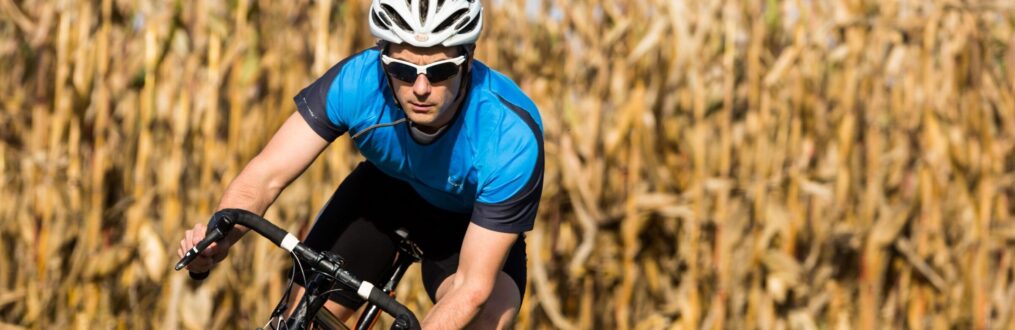 Fahrradfahrer mit Fahrradbrille und Fahrradhelm fährt vor einem Getreidefeld entlang.